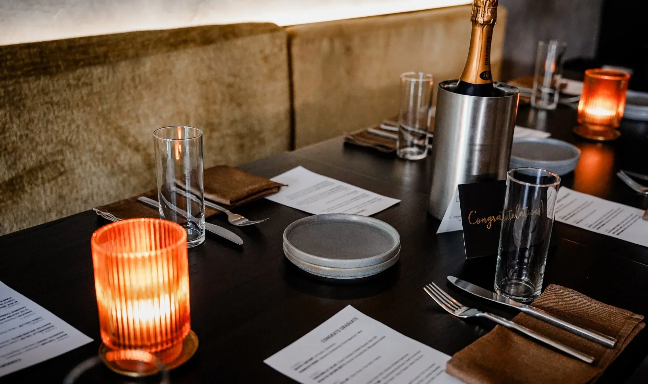 A booth set for a celebration with candles, a bottle of Champagne on ice, and printed menus