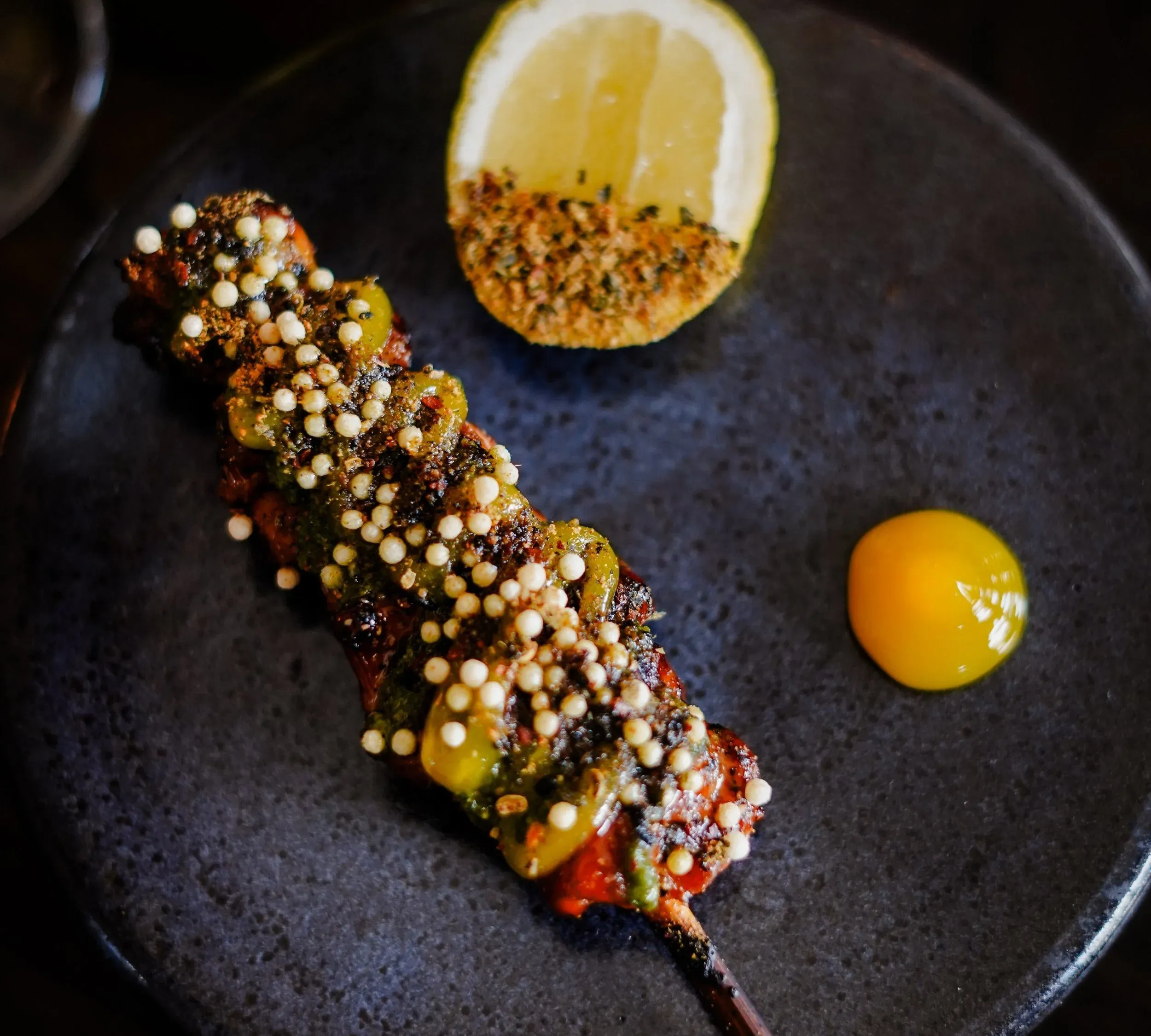 A grilled chicken skewer topped with crisp rice pearls and green sauce, with a charred lemon half